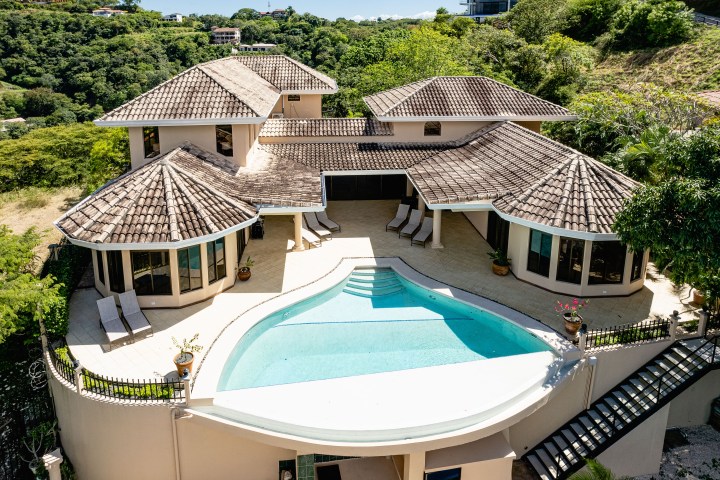 Aerial view of a house with curved pool, tiled roof, patio chairs, and surrounding greenery.
