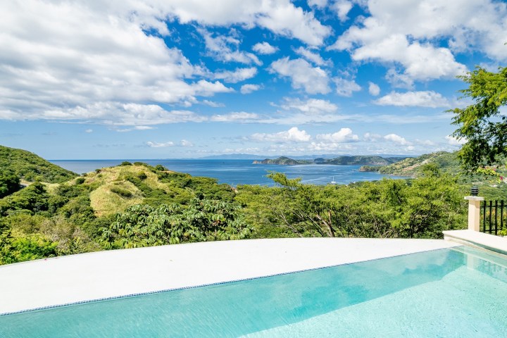 Infinity pool overlooking lush hills and a blue ocean under a partly cloudy sky.