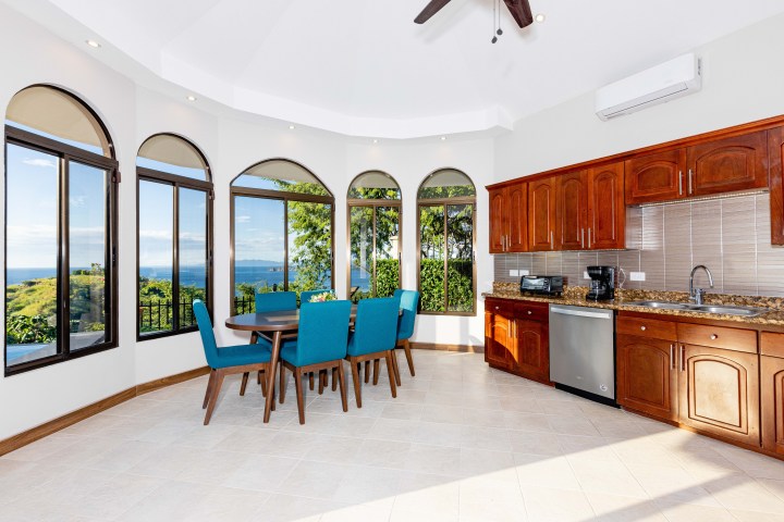 Bright kitchen and dining area with ocean view, wooden cabinets, and blue chairs.