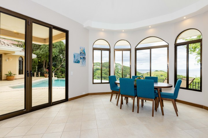 Dining room with blue chairs and large windows overlooking a pool and lush landscape outside.