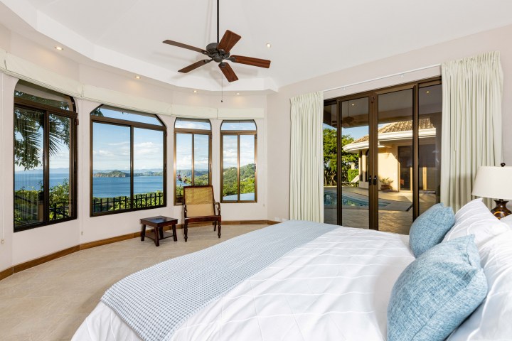 Bedroom with bay windows, ocean view, bed with blue pillows, and ceiling fan.