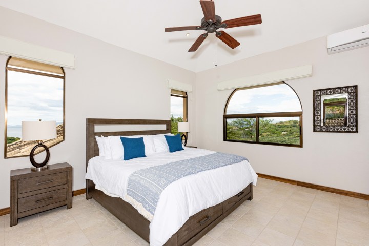 Bright bedroom with a king-size bed, blue pillows, ceiling fan, and large windows with scenic views.