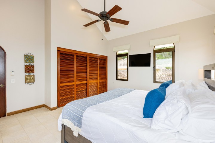 Modern bedroom with ceiling fan, wooden closet, and blue-accented bedding next to two windows and a TV.