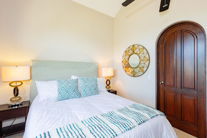Bedroom with white bedding, teal pillows, two lamps, and an arched wooden door next to a decorative mirror.