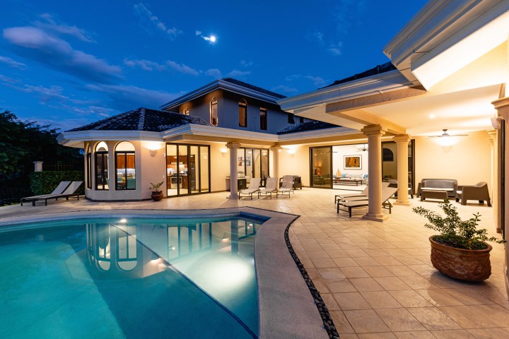 Luxury home with a lit pool and patio at dusk, featuring large windows and elegant outdoor seating.
