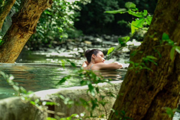 Person relaxing in a natural pool surrounded by lush forest, with trees framing the scene.