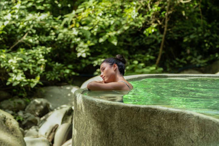 Woman relaxing in an outdoor stone pool surrounded by lush greenery.