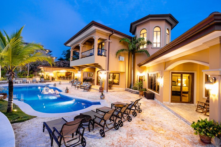 Luxurious villa exterior with pool, lounge chairs, and palm tree, illuminated at dusk.