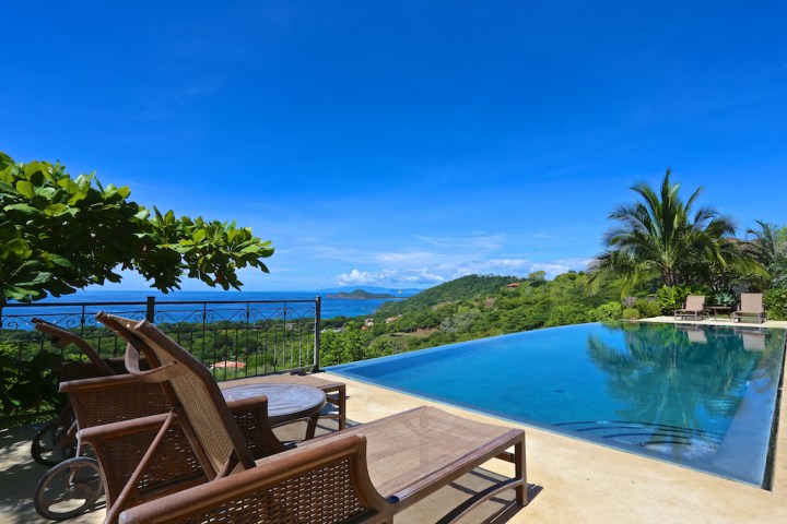 Infinity pool with lounge chairs overlooking ocean and hills under a clear blue sky.