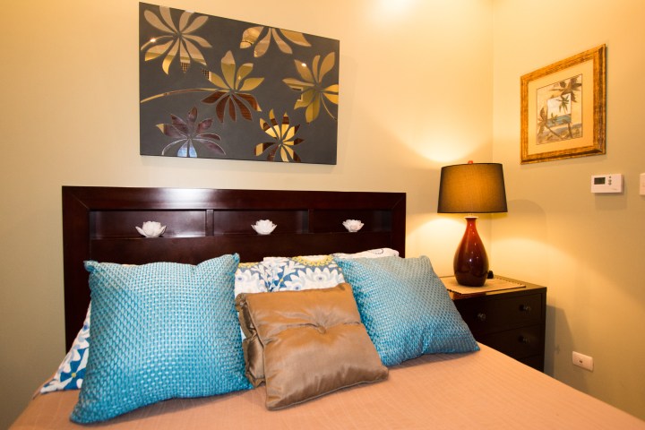 Cozy bedroom with blue pillows, floral artwork, and a bedside lamp on a wooden nightstand.