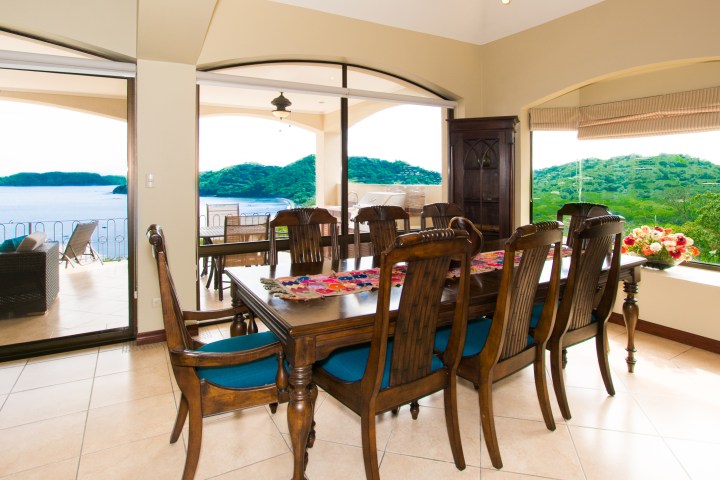 Dining room with wooden table, chairs, and ocean view from large windows.