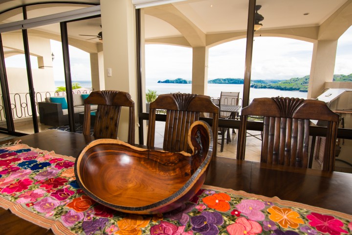 Room with wooden table, colorful runner, carved bowl, and ocean view through large windows.
