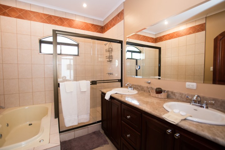 Bathroom with double sinks, shower, jacuzzi tub, and beige-tiled walls.