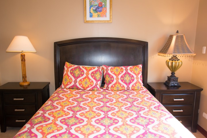 Bedroom with patterned bedspread, two lamps on dark wood nightstands, and a framed painting above the bed.