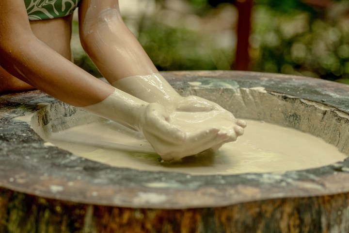 Person's hands covered in creamy substance, mixing it in a large circular wooden container.