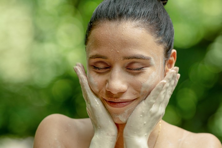 Woman with closed eyes applying face mask outdoors, hands on cheeks.