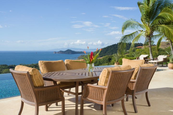 Outdoor patio set overlooking ocean with palm trees and hills in the background.