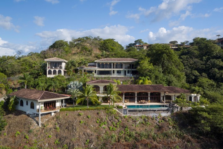 Luxury hillside villa with multiple levels, palm trees, and a pool surrounded by lush greenery.