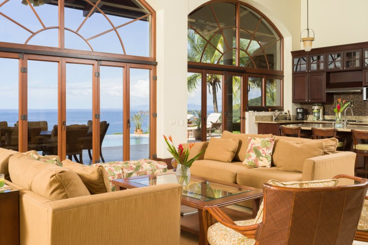 Spacious living room with ocean view, tan sofas, glass coffee table, and kitchen island with dark cabinets.