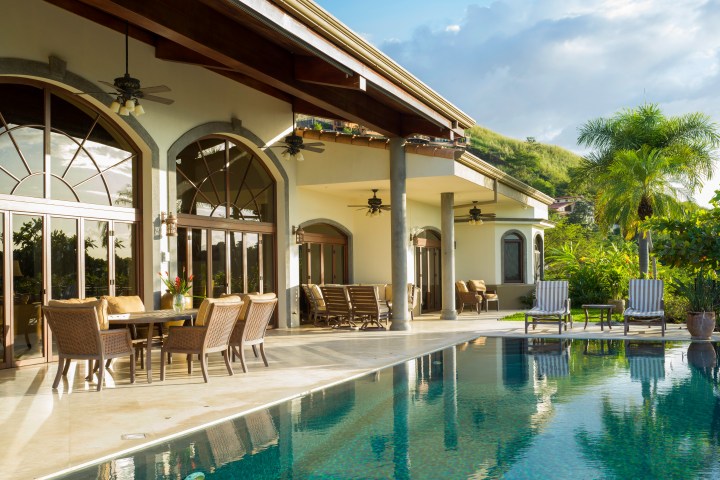 Luxurious house with arched windows and poolside seating in a tropical setting.