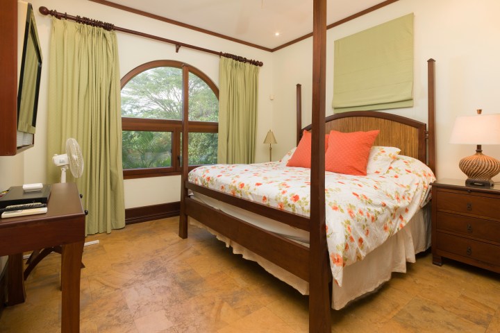 Cozy bedroom with a large bed, floral bedding, green curtains, wooden furniture, and a lamp on a nightstand.