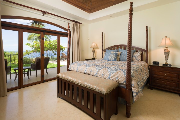 Luxurious bedroom with four-poster bed, ocean view through glass doors, and elegant wooden furniture.