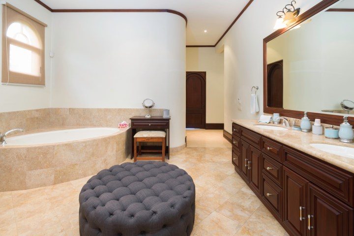 Luxurious bathroom with large mirror, double sinks, tub, and a cushioned ottoman.