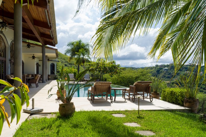 Poolside patio with chairs and table, tropical plants, and distant hills under a partly cloudy sky.