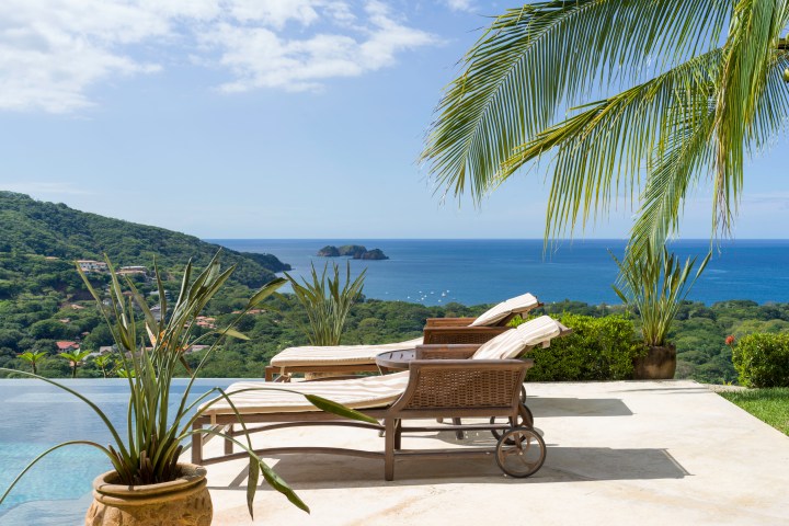 Two lounge chairs on a patio overlooking the ocean with palm trees and greenery.