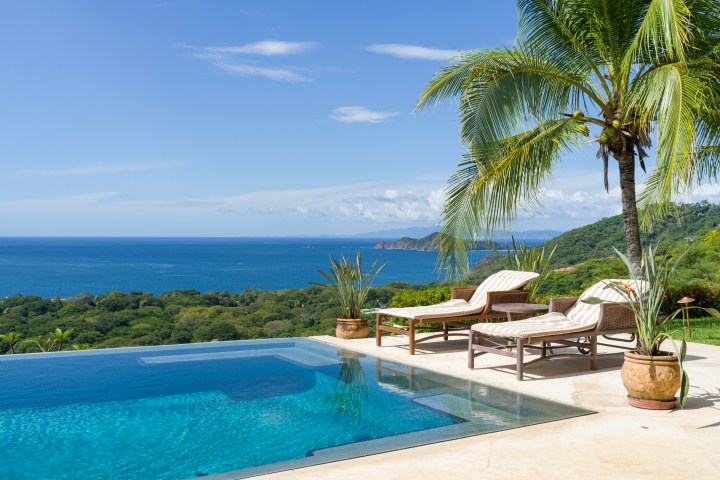 Infinity pool with loungers and palm trees overlooking ocean and green hillside.