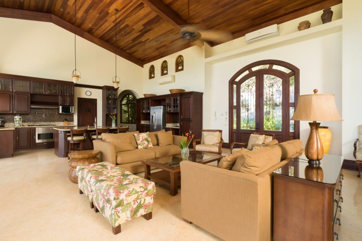 Spacious living room with brown furniture, high ceiling, large windows, and a modern kitchen in the background.