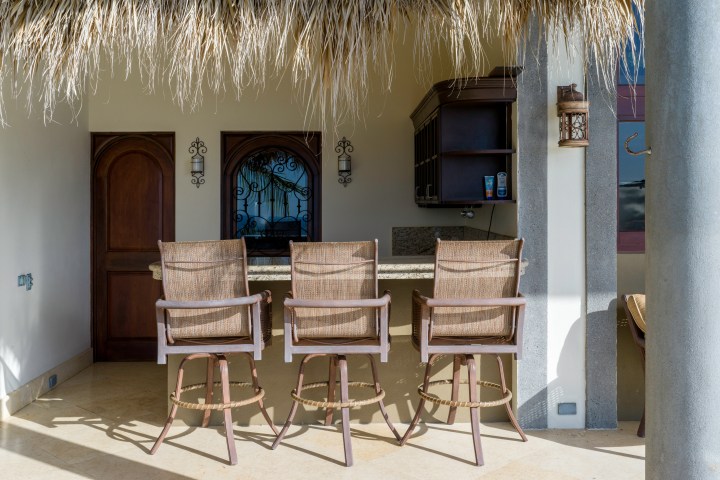 Three wicker bar stools at an outdoor bar with thatched roof and wooden doors.