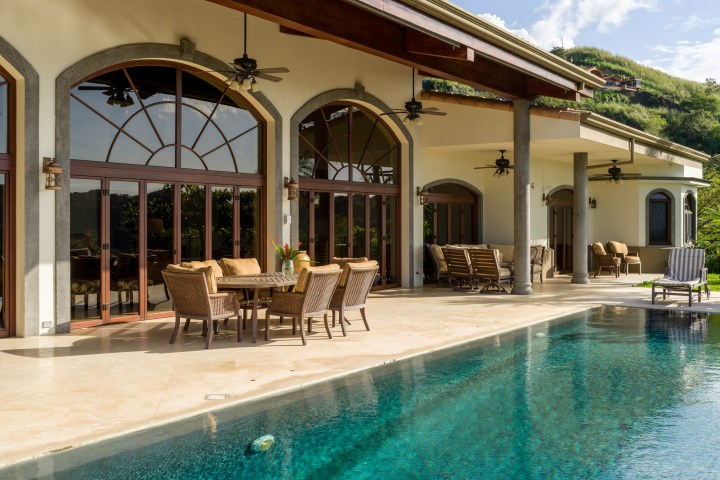 Luxury patio with wicker furniture, ceiling fans, and infinity pool next to large villa with arched windows.