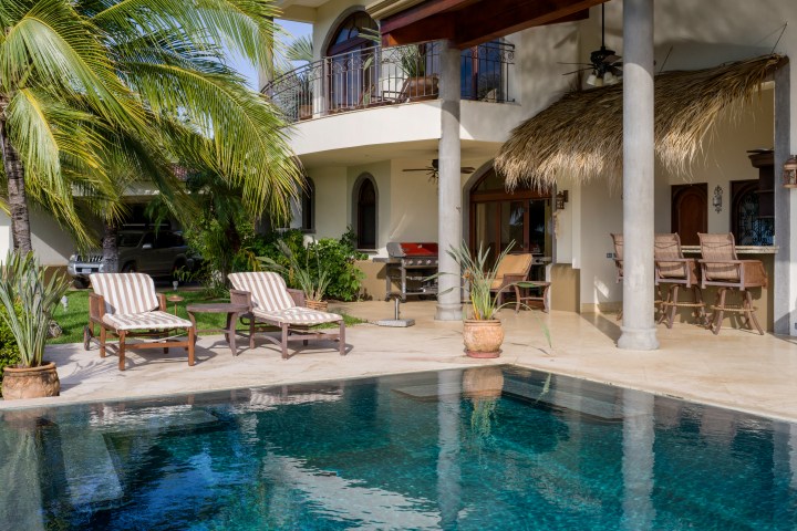 Tropical patio with pool, lounge chairs, and bar seating under a thatched roof near palm trees.