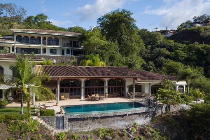 Luxury hillside villa with pool, surrounded by lush greenery and clear skies.