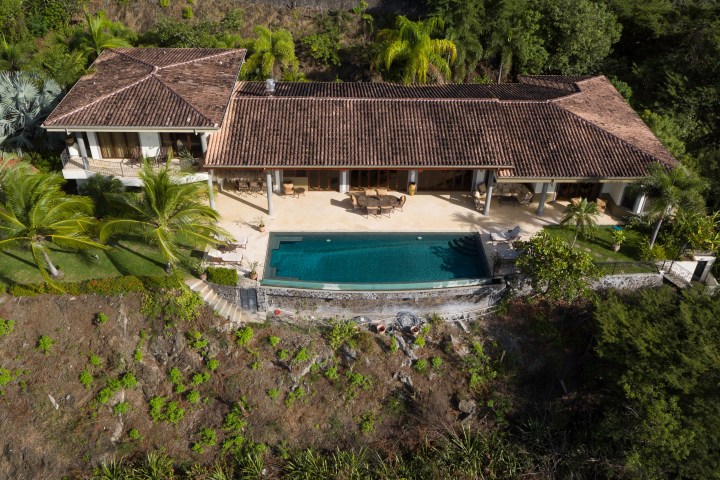 Aerial view of a house with a tiled roof and a rectangular swimming pool, surrounded by palm trees and greenery.