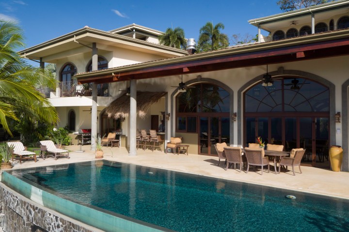 Luxury house with outdoor pool, palm trees, and patio dining area under a blue sky.