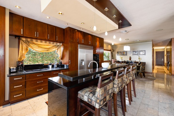 Modern kitchen with wooden cabinets, island, bar stools, pendant lights, and dining area in the background.