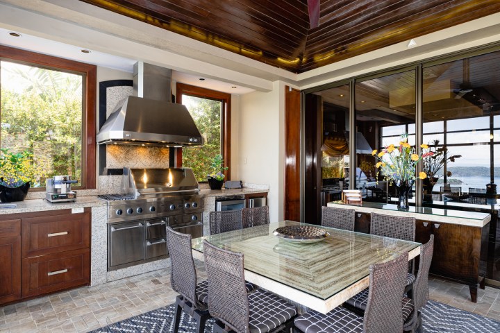 Modern kitchen with stainless steel appliances, glass dining table, wicker chairs, and large windows overlooking a view.