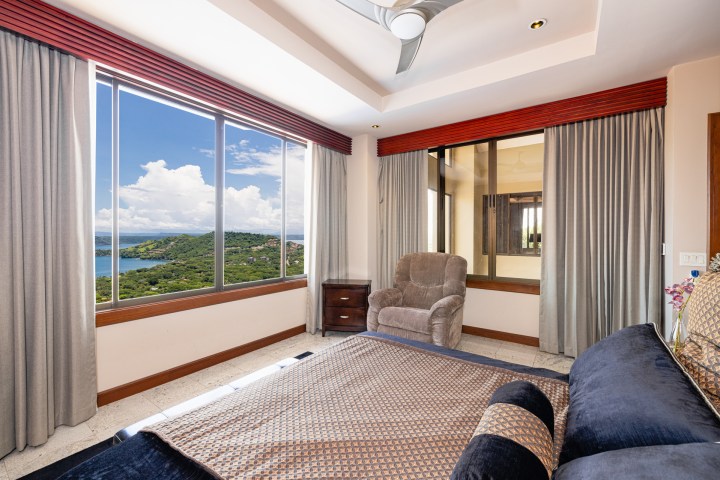 Bedroom with large windows, scenic view of hills and water, armchair, and bed.