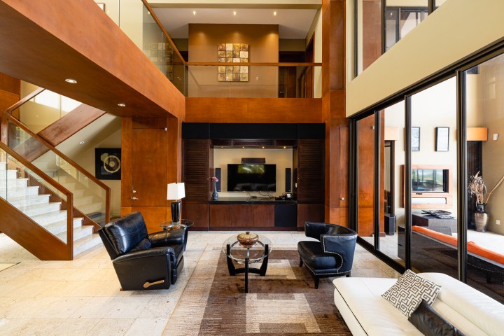 Modern living room with leather chairs, glass staircase, TV, and large windows.