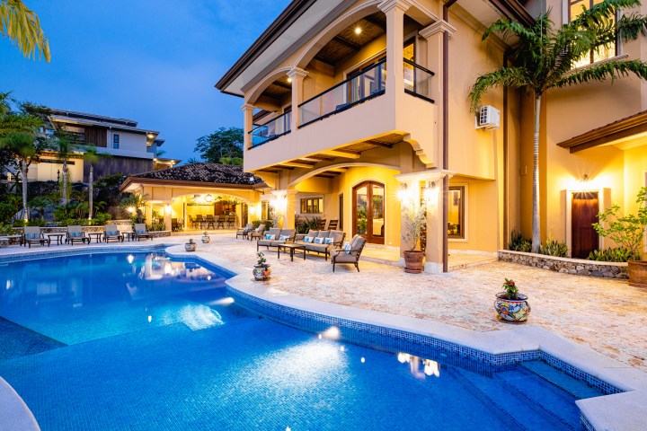Luxurious villa with a pool and patio seating, lit warmly at dusk with tropical plants surrounding.