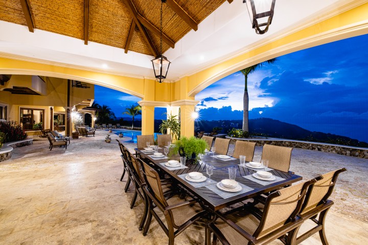Outdoor patio with dining table and chairs, view of pool and mountains at dusk.