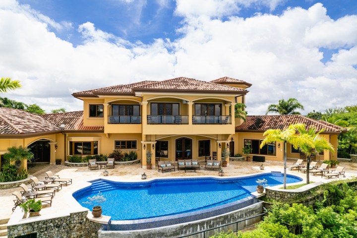Large villa with a curved pool, surrounded by tropical plants under a blue sky with clouds.