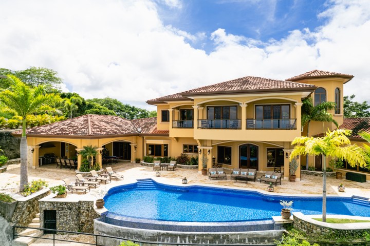 Luxury villa with pool, palm trees, and patio chairs under a blue sky.