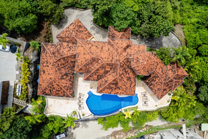 Aerial view of a house with a red-tiled roof and a blue pool surrounded by trees.