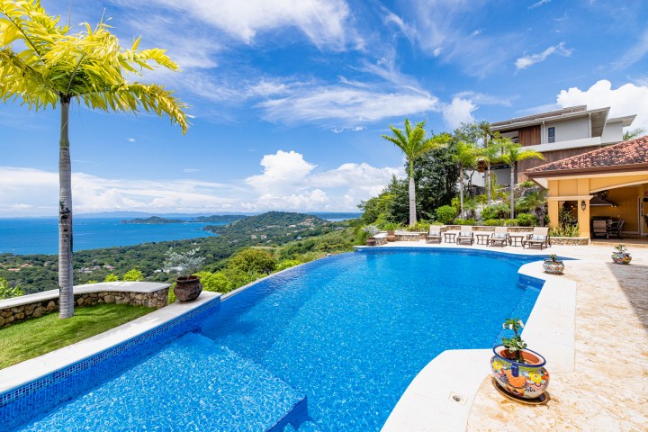 Infinity pool overlooking ocean and hills with palm trees and villa in background.