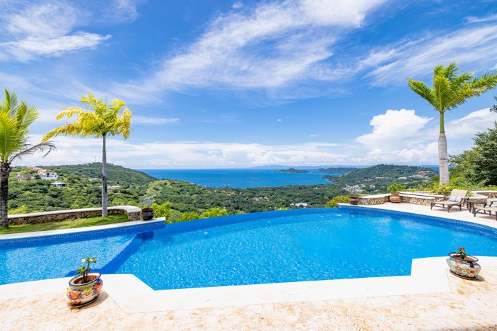 Infinity pool with a coastal view, surrounded by trees and decorative pots.