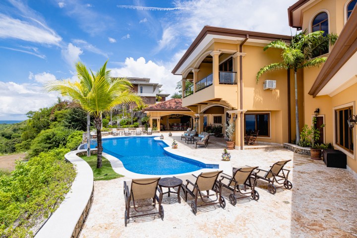 Luxurious villa with a pool, palm trees, and patio furniture under a blue sky.