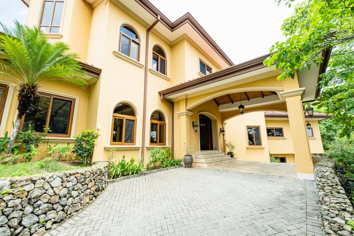 Two-story yellow house with arched windows, driveway, and lush green landscaping.